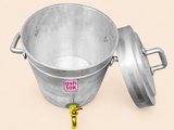 A high-angle view looking down into an open silver aluminum tea container on a light beige background. The image highlights the clean, empty metallic interior of the vessel, with its lid resting on the right side and a brass dispensing tap visible at the lower front. The Ashtok logo is printed on the container's exterior wall.