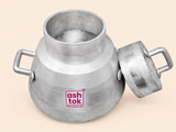 An angled top-down view of an open silver aluminum chai brewing pot on a neutral beige background. The pot has two sturdy side handles and a narrow circular opening showing its hollow interior. Its top lid with a metal handle rests leaning against the right side of the pot, and the magenta Ashtok logo is centered on the front.