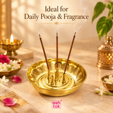 Traditional brass agarbatti stand displayed in a warm festive pooja setting with three burning incense sticks releasing gentle smoke. The polished golden incense holder features a round ash-catching bowl and five upright incense tubes, surrounded by jasmine flowers, rose petals, brass pooja accessories and soft decorative lighting. Suitable for daily prayers, meditation, spiritual rituals and home mandir arrangements.