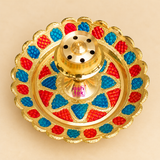 Top view of a traditional Brass Incense Stick Holder featuring a circular golden brass tray with an ornamental scalloped edge and alternating red and blue Meenakari-style decorative patterns. The centre features a raised brass incense holder with multiple circular openings arranged around a cone-shaped top. The symmetrical design highlights the detailed floral arrangement, decorative metalwork and colourful surface finish of the 3.5-inch-diameter brass agarbatti stand, suitable for pooja rooms, home temples