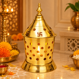 Traditional Brass Akhand Diya illuminated with a warm golden flame inside its decorative perforated brass enclosure, displayed in an elegant home pooja setting with marigold flowers, brass pooja accessories, and soft festive lighting. The polished brass Akhand Jyoti lamp features a conical Swastik-design cover, ornamental pointed finial, round ventilation holes, and a wide stepped base. The glowing flame is visible through the circular openings, creating a devotional atmosphere suitable for Navratri, Diwali
