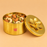 Decorative brass coloured metal dry fruit gift box displayed open on a warm beige festive tabletop and filled with almonds, cashews, pistachios and golden raisins. Its polished golden circular lid with a raised flower-shaped topper rests beside the storage container. The arrangement showcases its use for presenting dry fruits during Diwali celebrations, wedding functions, festive gatherings and traditional household gifting.