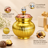 Decorative brass and copper gift box shown with its original retail packaging on a premium beige studio background, featuring a rounded ribbed golden brass body, polished copper-toned lid, crystal-style faceted knob and compact pedestal base, presented beside the matching printed gift box to show the complete product presentation, suitable for storing chocolates, dry fruits, sweets, small gifts and festive treats for weddings, housewarming occasions, anniversaries, Diwali gifting, family celebrations and tr
