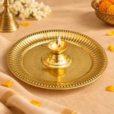 Traditional brass diya with attached pooja thali displayed in a festive prayer setting with a glowing flame in the central oil lamp. The polished golden brass plate features decorative engraved patterns, a raised beaded border and a centrally mounted diya cup. Surrounded by jasmine flowers, marigold petals and traditional pooja accessories, the image illustrates its use during Diwali celebrations, daily prayers, aarti rituals and home mandir worship.
