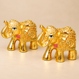 Front angled product view showing a pair of golden Brass Elephant Idols with raised trunks, elaborate head ornaments and decorative traditional carvings. The two miniature elephant figurines are positioned facing one another against a neutral beige background, clearly displaying their curved trunks, sculpted ears, ornamental body patterns, polished golden finish and four sturdy legs. Each statue measures 2 inches in length, 1 inch in width and 1.5 inches in height. Suitable for home temple decoration, offic