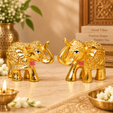 Set of 2 golden Brass Elephant Idols displayed facing each other in an elegant traditional home decoration setting with a warm beige background, decorative brass diya, white flowers and ornamental accessories. Both miniature elephant figurines feature raised trunks, polished golden surfaces, intricate embossed body patterns and detailed ornamental head designs. Each elephant measures 2 inches in length, 1 inch in width and 1.5 inches in height. Suitable for home entrance decoration, living room shelves, off