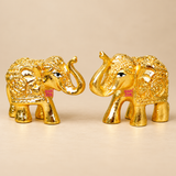 Set of 2 decorative Brass Elephant Idols featuring a polished golden finish, beautifully raised trunks, intricate embossed traditional patterns and ornamental detailing across their bodies. Each miniature elephant figurine measures 2 inches in length, 1 inch in width and 1.5 inches in height. The two elephants are displayed facing one another on a clean beige studio background, highlighting their detailed head ornaments, expressive eyes, curved trunks and sturdy four-legged design. Ideal for traditional hom