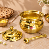Traditional brass ghee pot shown in a warm lifestyle dining setting filled with golden clarified butter or ghee and styled alongside Indian flatbreads and serving dishes. The matching lid and brass serving spoon are placed beside the pot to demonstrate practical everyday use. The polished golden container features a rounded traditional body and wide rim, making it suitable for serving ghee with roti, chapati, paratha, rice and other Indian meals.