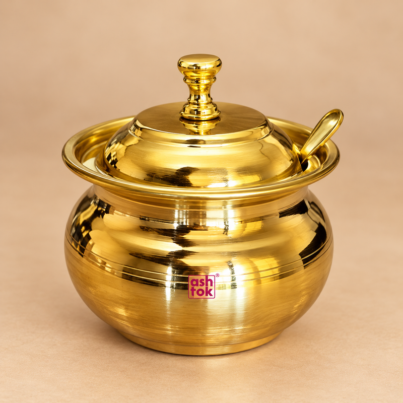 Premium brass ghee pot with matching fitted lid and serving spoon displayed as a single traditional ghee serving container on a clean warm beige studio background. The pot features a rounded traditional handi-style body, wide rolled rim, polished golden brass finish, matching domed lid with decorative knob and a dedicated spoon positioned neatly through the side opening. 