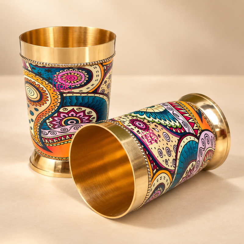 Brass glasses with printed design premium drinkware for special occasions featuring two elegant brass tumblers with a polished golden finish and vibrant multicolor paisley-inspired artwork. One glass is upright and one is placed sideways to show the shape, finish and interior, professionally presented on a premium beige studio background for gifting, festive serving and decorative drinkware use.