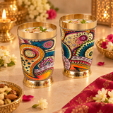 Brass glasses with printed design shown in a festive lifestyle setting filled with a refreshing traditional beverage and surrounded by flowers, decorative bowls and celebratory table styling. The vibrant printed brass drinkware is ideal for serving guests during festivals, housewarming events, family gatherings, special occasions and traditional celebrations.