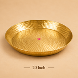 Product photograph showcasing an extra-large 20-inch brass puja tray with a reflective hammered brass finish. Placed on a beige backdrop with '20 Inch' written below in brown serif typography and the pink Ashtok logo printed in the middle.