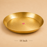 Product image of a large 18-inch circular brass puja thali with a traditional handcrafted hammered texture and smooth raised rim. Resting on a solid beige surface with text reading '18 Inch' centered underneath and the Ashtok logo in the center.