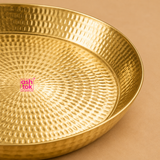 Macro close-up photograph of a handcrafted brass puja plate highlighting the detailed concentric hammered texture across its surface and smooth sloping raised side rim, set against a soft beige background with the Ashtok brand logo centered.
