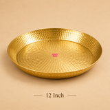 High-angle product photograph of a round handcrafted brass puja thali with a hammered textured finish set against a plain beige background. Text below the tray reads '12 Inch'. A subtle pink 'ashtok' brand watermark logo is placed in the center of the brass plate
