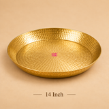 High-angle product photograph displaying a round 14-inch handcrafted brass thali plate with a dimpled hammered finish resting on a beige background. Text centered below reads '14 Inch', with a magenta Ashtok brand logo watermark printed in the middle of the thali surface.