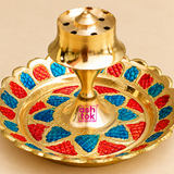 Close-up product photograph of a decorative Brass Incense Stick Holder showing its polished golden brass central agarbatti stand, circular incense holding holes, raised pedestal support and detailed red and blue Meenakari-style decorative patterns. The surrounding brass tray features a scalloped floral border, textured coloured sections and reflective golden finishing. Measuring 3.5 inches in diameter and 2 inches in height, this traditional incense holder is suitable for home temple accessories, daily pooj