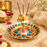 Decorative Brass Incense Stick Holder displayed with three burning agarbatti sticks releasing gentle incense smoke in a traditional pooja setting. The polished golden brass holder features a raised central incense stand, multiple agarbatti holes and a circular tray decorated with vibrant red and blue Meenakari-style patterns. Surrounded by white flowers, rose petals and warm festive lighting, the 3.5-inch-diameter and 2-inch-high incense holder is suitable for daily prayers, home mandir decoration, meditati