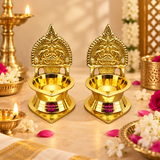 Lifestyle image of a Brass Kamakshi Deepam pair placed in a festive pooja-inspired setting. The two polished brass deepams are arranged on a warm beige surface surrounded by flowers, petals, and traditional décor elements that create a devotional atmosphere. The image helps customers visualize how the lamps can be used in a home temple, during pooja rituals, festive celebrations, and as elegant traditional décor pieces for spiritual spaces.