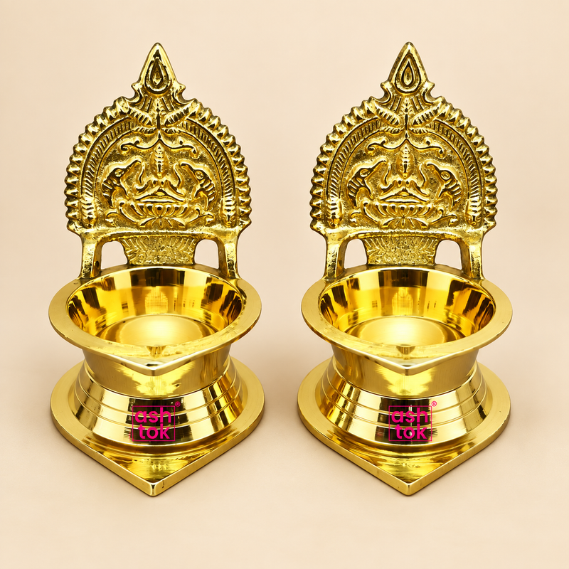 Pair of polished brass Kamakshi deepam lamps displayed on a plain beige studio background. The image shows two identical traditional brass deepams placed side by side, each featuring a decorative backplate with intricate temple-style detailing, a deep oil bowl in front, and a sturdy round stepped base. The finish is bright and reflective, highlighting the clean craftsmanship and classic devotional design. Suitable for pooja use, home temple décor, festive gifting, and traditional occasions.