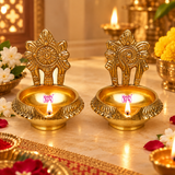 Set of 2 Brass Kuber Diyas displayed in a festive pooja setting with lit flames, flowers and warm decorative background. The golden brass diyas feature a detailed floral backplate and traditional bowl shape, making them suitable for daily pooja, Diwali decoration, home temple use and festive celebrations.