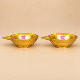 Side-profile product photograph of two Brass Kuber Diyas featuring a traditional shallow brass oil bowl structure, polished golden metallic finish, pointed wick spouts, engraved geometric detailing around the outer surfaces, and compact circular pedestal bases. The side-angle presentation clearly displays the gently curved bowl shape