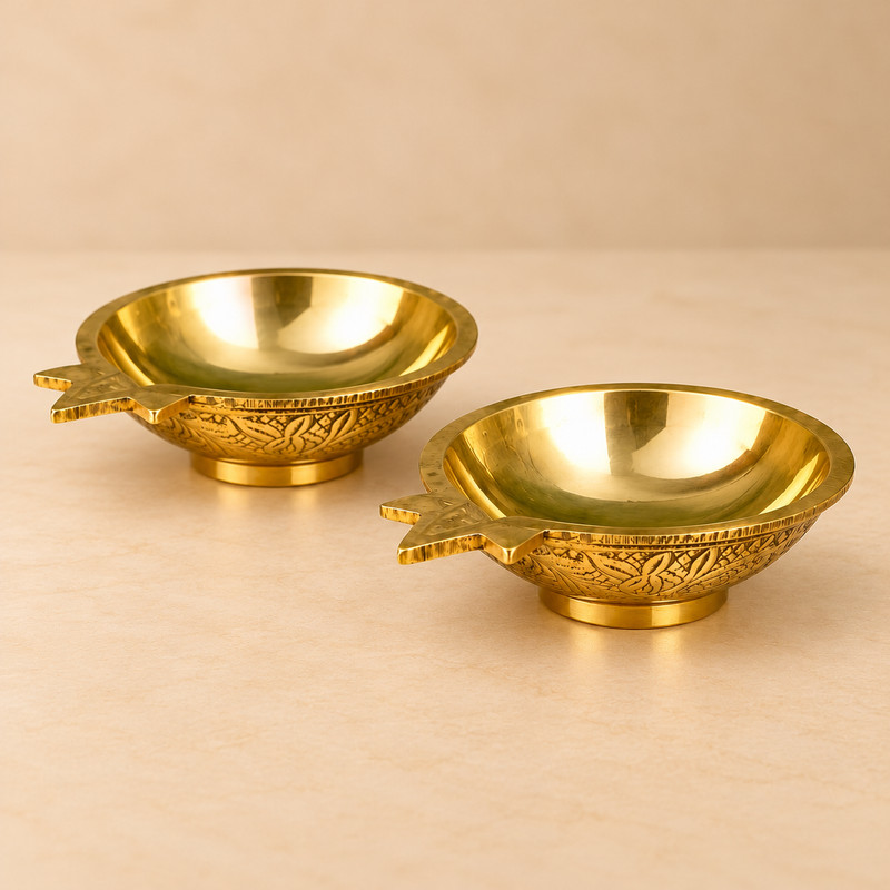 Three-quarter angle photograph of two traditional Brass Kuber Diyas featuring polished golden brass interiors, wide circular bowl openings, distinctive pointed wick spouts extending from one side, beautifully engraved floral and leaf patterns around the outer bowl surfaces, and stable raised circular bases. The detailed perspective showcases the smooth reflective brass finish and compact proportions of these traditional pooja oil lamps. Each diya measures 3 inches in length, 2.5 inches in width, and 1 inch 
