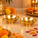 Two traditional Brass Kuber Diyas illuminated with warm golden flames in an elegant festive pooja setting, surrounded by fresh marigold flowers, decorative brass pooja accessories, and colourful rangoli arrangements. Each diya features a shallow polished brass oil bowl, distinctive pointed wick spout, intricate engraved floral detailing, and a stable circular pedestal base. The warm glowing flames complement the rich golden brass finish, creating a devotional atmosphere suitable for Diwali celebrations, Dha