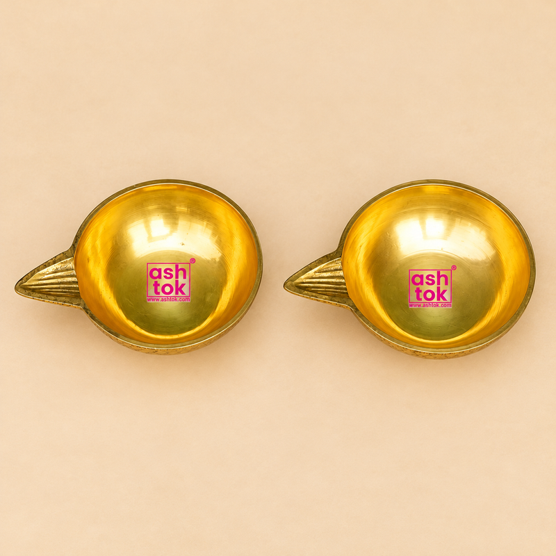 Detailed top-view photograph of Brass Kuber Diya Set of 2 Pcs showcasing two identical traditional golden brass oil lamps with smooth circular bowl-shaped interiors, broad polished rims, and decorative pointed wick spouts extending outward from one side of each diya. The elevated perspective highlights the symmetrical oil reservoirs, reflective golden brass surfaces, and distinct tapered spout design.