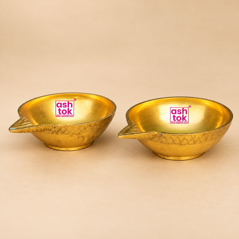 Brass Kuber Diya Set of 2 Pcs featuring two matching traditional brass pooja oil lamps with a polished golden finish, smooth rounded oil-holding bowls, beautifully designed pointed wick spouts extending from one side, intricate decorative geometric engravings around the outer rims, and small stable circular bases. Each brass diya measures 3 inches in length, 2.5 inches in width, and 1 inch in height. The two compact brass oil lamps are displayed side by side against a premium beige studio background.