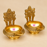 Top and angled view of Brass Kuber Diya set of 2 showing the round brass bowl, pointed front spout, decorative outer border and upright floral backplate. These traditional brass diyas are compact in size and suitable for pooja setups, festival decoration and home temple display.
