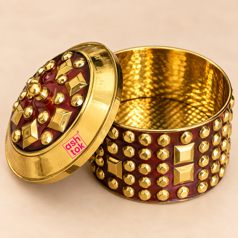 Close-up image of Brass KumKum Box with open lid showing the polished golden interior, maroon body with brass stud work and Ashtok logo on a beige studio background.