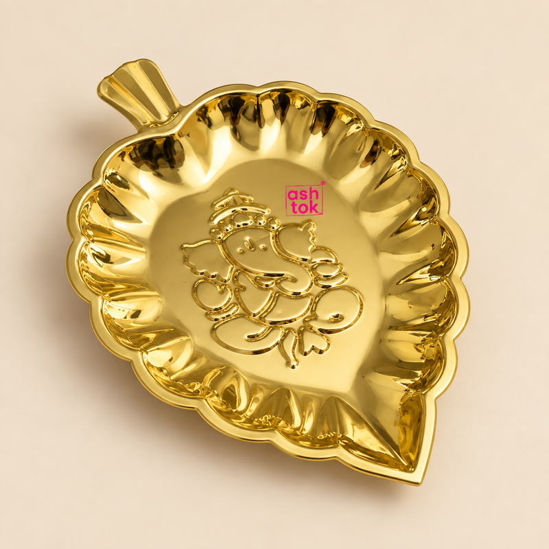 Brass leaf tray with a polished golden finish, decorative leaf-shaped form, scalloped raised rim and embossed Ganesh design in the center, professionally presented on a premium beige studio background for puja, festive decor and gifting.