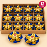 Pack of 12 decorative brass lotus diyas featuring polished golden brass oil bowls, central wick holders, curved lotus-shaped petals and vibrant royal blue floral bases. The diyas are arranged in individual compartments inside a simple brown cardboard box, with additional display pieces shown in front. Ideal for Diwali celebrations, traditional puja ceremonies, wedding return gifts, festive decorations and gifting in larger quantities.