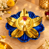 Traditional golden brass lotus diya with a vibrant royal blue floral base displayed with a glowing flame on an elegant beige marble surface. Surrounded by jasmine flowers, yellow marigolds, rose petals and warm decorative lighting, this beautifully designed oil lamp creates a festive atmosphere suitable for Diwali celebrations, daily worship, traditional home puja arrangements and decorative festive displays.