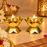 Brass Lotus Diya Set of 2 Pcs beautifully illuminated with warm golden flames in an elegant traditional pooja setting, surrounded by fresh white flowers, decorative brass accessories, and soft festive lighting. Each golden brass diya features a gracefully shaped lotus-petal bowl, smooth circular oil reservoir, raised central wick holder, and sturdy lotus-shaped pedestal base. The two glowing brass oil lamps create a peaceful devotional atmosphere suitable for home mandir worship, Diwali celebrations, Lakshm
