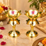 Two traditional Brass Lotus Diya Stands beautifully displayed with glowing oil lamp flames in a festive home decoration setting. Each golden brass diya measures 5.5 inches in height and 2.5 inches in diameter, featuring an open lotus-shaped bowl, central wick holder, decorative layered petals and round pedestal base. Surrounded by marigold flowers, jasmine blossoms, rose petals and warm decorative lighting, these lotus oil lamps are ideal for Diwali celebrations, daily pooja, festive rituals, housewarming c