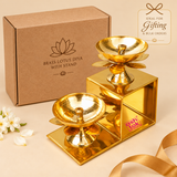 Decorative Brass Lotus Diya with Stand presented alongside simple cardboard packaging on a premium beige studio background. The polished golden brass product features a geometric two-step stand holding two lotus-shaped oil lamps at different heights, each with a smooth circular bowl, central wick holder and ornamental lotus petal detailing. This traditional brass diya stand is suitable for Diwali gifts, housewarming presents, festive gifting, wedding gifts, decorative home accessories and bulk gift orders. 