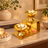 Brass Lotus Diya with Stand displayed as an elegant decorative lighting accessory in a modern home interior with a warm beige colour palette, minimalist ceramic vase, white flowers and contemporary tabletop decorations. The polished golden brass two-step stand features two traditional lotus-shaped oil lamps with glowing flames positioned at different heights. Each diya has a smooth round bowl, central wick holder and decorative lotus petals underneath. The geometric brass structure complements modern living