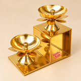 side angled view of the Brass Lotus Diya with Stand featuring two polished golden brass oil lamps mounted on a decorative two-step geometric frame. The elevated perspective clearly shows the wide circular diya bowls, central wick holders, smooth reflective brass interiors and lotus-shaped petals beneath each holder. The arrangement features one diya at the lower level and another at the upper level, supported by a rectangular metal structure with a bright golden finish. Ideal for displaying the detailed.