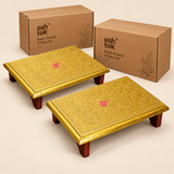 Two-piece Brass Marriage Chowki set displayed against a clean beige studio background with simple brown cardboard packaging boxes. Both rectangular platforms feature richly embossed golden brass sheet surfaces decorated with traditional floral and vine patterns, detailed ornamental borders, polished golden edges and four dark brown wooden supporting legs. The two matching chowkis are arranged separately to showcase their traditional craftsmanship and coordinated design.