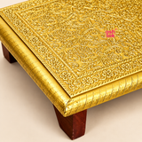 Close-up product photograph of a traditional Brass Marriage Chowki highlighting the intricate embossed floral patterns, decorative vine motifs, ornamental border designs and beautifully finished golden brass sheet surface. The enlarged view reveals fine traditional detailing across the rectangular platform, along with the polished golden corner, engraved edge decorations and sturdy dark brown wooden supporting leg. Soft beige studio lighting enhances the detailed.