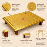 Detailed features image of a traditional Brass Marriage Chowki highlighting its handcrafted golden brass sheet covering, intricate embossed floral and vine patterns, decorative engraved borders, sturdy wooden legs and low rectangular platform design. Close-up illustrations showcase the fine ornamental workmanship across the brass surface, polished golden edges and dark brown wooden supports. Presented against a warm beige studio background with informative feature callouts, demonstrating the traditional.
