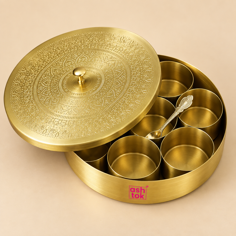 Traditional round brass masala box with seven removable inner spice bowls, matching decorative lid and serving spoon displayed on a clean warm beige studio background. The spice storage box features a smooth brushed golden brass outer body, seven individual cylindrical containers arranged neatly inside, an intricately engraved circular lid with a central knob and a decorative metal spoon placed within the set.