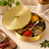 Traditional brass masala box shown in a warm kitchen lifestyle setting with its seven inner bowls filled with colorful Indian spices and whole ingredients including turmeric powder, red chilli powder, cumin seeds, black pepper, coriander seeds and dried red chillies. The decorative engraved lid is partially open and the matching serving spoon is positioned inside the box, demonstrating practical everyday kitchen use. 