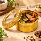 Traditional brass masala storage box displayed on a warm beige kitchen countertop with its transparent viewing lid removed and placed beside the container. The round golden brass box is filled with whole Indian spices including green cardamom, cinnamon sticks, star anise, bay leaves, dried red chillies and black peppercorns. The lifestyle image demonstrates its use for kitchen spice storage and organization.