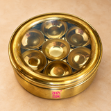 Brass masala box with transparent glass lid and seven inner spice bowls, featuring a polished golden finish and round compact design, presented on a premium beige studio background for kitchen spice storage.