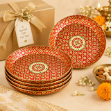 Festive gifting arrangement featuring a stack of four red and gold Meenakari brass trays next to a prop tray, a wrapped gold gift box tied with a satin bow and tag reading 'A Special Gift For You', a brass bowl of dry fruits, and marigold flowers.
