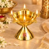 Traditional brass Panch Batti Diya shown lit with five cotton wicks in a warm festive pooja setting. The five flames are arranged around the lotus-inspired brass bowl, demonstrating the intended use of the deepam during prayer and ceremonial lighting. The polished golden lamp is styled with subtle flowers, brass décor and warm beige surroundings that create a devotional festive atmosphere without distracting from the product. This image demonstrates how the five-wick brass diya can be used for daily pooja.