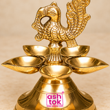 Detailed close-up view of a traditional Brass Panchmukhi Peacock Design Diya highlighting the intricately sculpted golden brass peacock figure, decorative feather patterns, five surrounding oil lamp bowls, and finely polished metal finish. The ornamental peacock finial features a gracefully curved neck, carefully shaped head and eye, distinctive beak, layered wing feathers, and an elaborate upright tail decorated with raised patterns and textured carvings. The detailed bird figure is supported by a short fl
